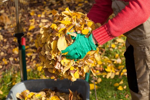 A person wearing bright green gloves and a red long-sleeve sweater is holding a large, tightly-packed bundle of yellow, brown, and orange fallen autumn leaves. The bundle appears freshly collected and is being prepared for disposal or removal. In the background, there is a garden area with scattered leaves on the ground, and a wheelbarrow is positioned nearby, partially visible at the bottom of the image. The scene is outdoors, likely in a residential garden or park setting, with natural lighting highlighting the vibrant colors of the leaves. This image reflects the process of garden clearance or leaf collection, which Garden Clearance Ickenham, a company specialising in rubbish removal services, often facilitates for clients in the Ickenham area.