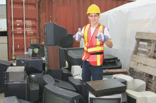 A male worker wearing a yellow safety helmet, high-visibility orange and yellow vest, and work gloves, stands outdoors in front of a pile of discarded electronic devices and office equipment, including old monitors, keyboards, and other small electronic debris. The background features a large brown shipping container with some text and a white sheet or tarpaulin cover on one side, indicating a waste collection or clearance area in a commercial or industrial yard. The worker is smiling and giving a thumbs-up gesture, conveying confidence in rubbish removal services. The scene suggests an organized effort to clear household or office waste, with a focus on electronic waste, in an environment associated with rubbish clearance in the Ickenham area. The lighting is natural, and the overall setting emphasizes a professional approach to waste disposal, aligning with Garden Clearance Ickenham’s rubbish removal services.