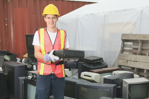A young man wearing a yellow safety helmet and a high-visibility pink and yellow vest, smiling while holding a black electronic device, standing outdoors in front of a collection of discarded electronic equipment such as old computer monitors and printers. The background includes a red wooden fence and a white tarp-covered structure, suggesting a waste disposal or recycling site. This scene illustrates rubbish removal and electronic waste collection services, with the individual representing a professional from a company like Garden Clearance Ickenham. The surrounding environment contains various obsolete electronic items stacked and lined up on a paved surface, indicative of a responsible disposal or clearance process. The lighting is natural, and the overall setting is clean, organized, and utilitarian, emphasizing the importance of proper rubbish removal for electronic waste in the local area near Ickenham.