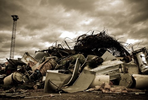 A large pile of discarded metal, plastic, and debris sits on an outdoor surface, with various items including vehicle parts, broken appliances, and twisted metal sheets. The materials vary in color, primarily shades of grey, white, and metallic, with some rusted and weathered textures visible. Behind the pile, a dense amount of tangled branches and foliage is partially visible on top of the debris, creating an uneven and chaotic heap. In the background, an overcast sky with dark clouds contributes to a moody atmosphere, and a tall industrial crane or tower is visible on the left side of the image. The scene suggests an environment where rubbish clearance services might be required, possibly in a location near Ickenham, with the debris forming part of an industrial or renovation waste collection. The focus is on the disorganized and cluttered nature of the waste, highlighting the need for professional rubbish removal, such as that provided by Garden Clearance Ickenham.