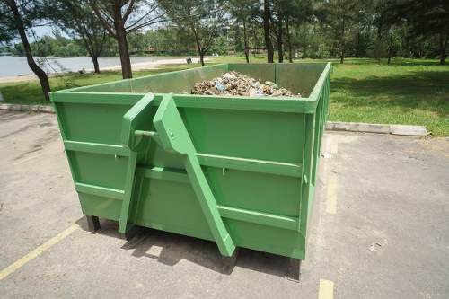 A large, rectangular green skip bin filled with a mixture of garden debris including soil, twigs, and organic waste, positioned on a paved area adjacent to a grassy park with trees and a body of water in the background. The skip bin is made of sturdy metal with horizontal reinforcement bars, and the surrounding environment suggests a typical outdoor setting in Ickenham. The scene captures the bin ready for rubbish removal, reflecting services provided by Garden Clearance Ickenham for garden waste disposal. Natural daylight illuminates the scene, highlighting the texture of the debris and the vibrant green of the bin, with a curb and parking area in the foreground and a lush, open space with mature trees and calm water in the background.