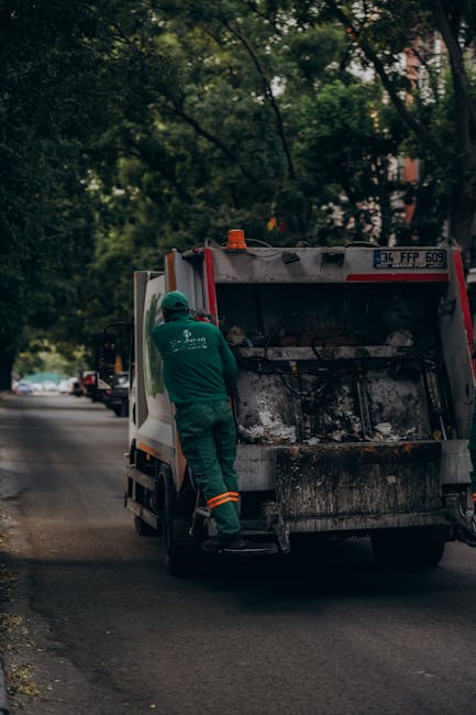 Two waste collection operatives dressed in high-visibility orange and red jackets with reflective strips are walking on a city street and pushing a wheeled trolley loaded with waste bins. One operativ