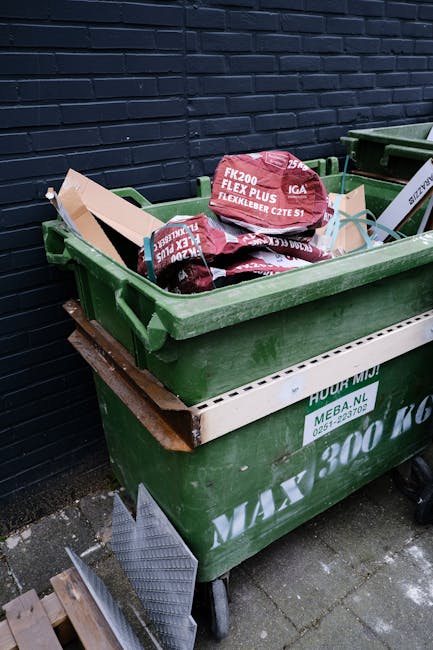 A large green wheelie bin filled with bags of gardening or building materials, including several red and black bags labeled 'FLEXLEADER CZE S1' and 'FK200 FLEXY PLUS', is positioned on a paved area ag